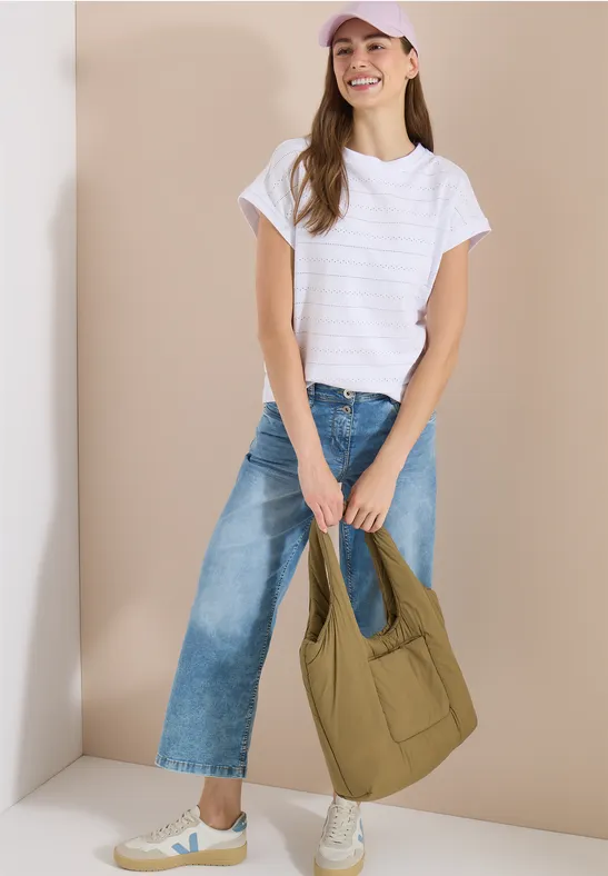 Frau trägt weißes T-Shirt, weite Jeans, weiße Sneaker und hält eine große olivgrüne Umhängetasche.