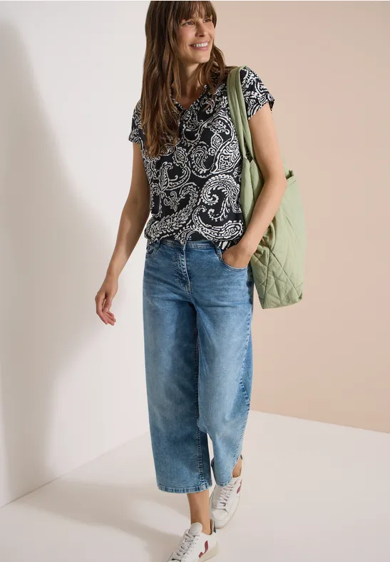Vrouw met een gemusterd t-shirt, wijde cropped jeans, witte sneakers en een groen doorgestikt tasje.