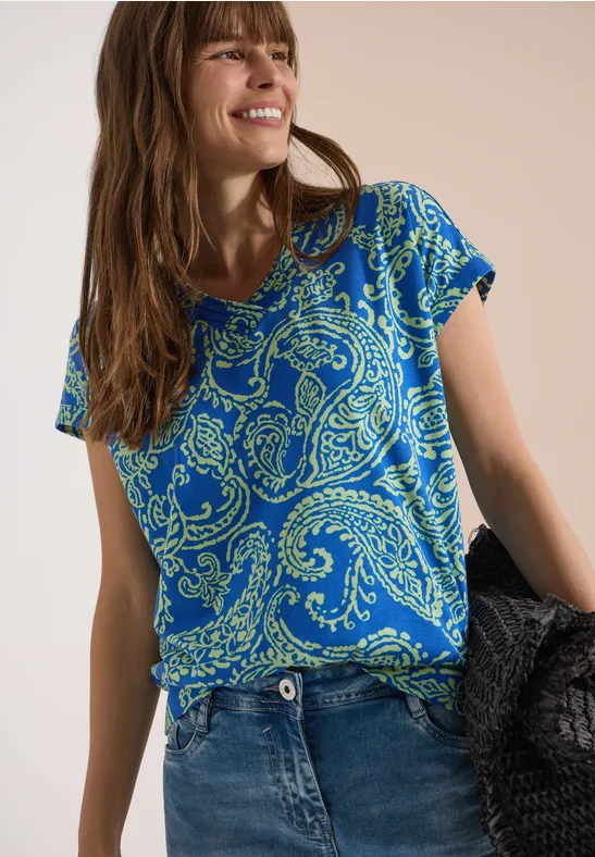 Vrouw draagt een blauw paisley-T-shirt en jeans en houdt een zwarte tas voor een neutrale achtergrond.