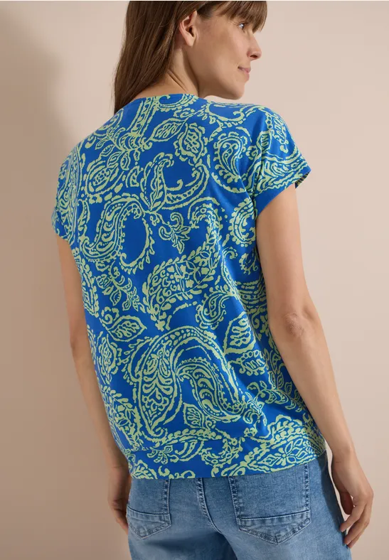 Vrouw van achteren in een blauw t-shirt met groene paisleymotieven en een spijkerbroek.