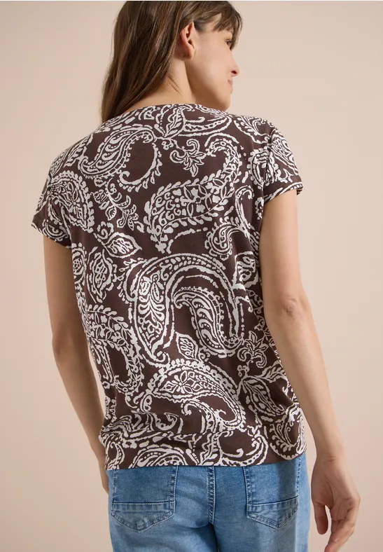 Vrouw van achteren in een bruin-wit paisley T-shirt en een blauwe spijkerbroek.