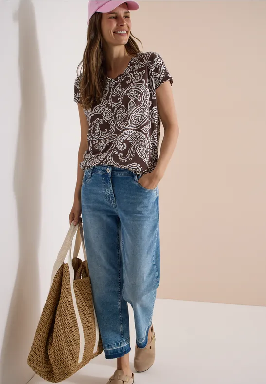 Vrouw in gemusterd shirt, blauwe jeans en grote rieten tas, casual look met loafers.