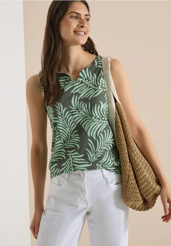 Vrouw in een groen topje met bladprint, witte broek en een gevlochten strozak.