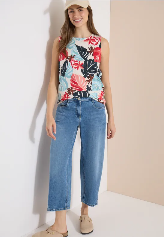 Frau in floralem ärmellosem Top, weiten blauen Culotte-Jeans und beigefarbenen Slippern vor hellem Hintergrund