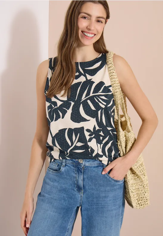 Frau in weißem Top mit schwarzem Blättermuster, blauen Jeans und geflochtener Schultertasche.