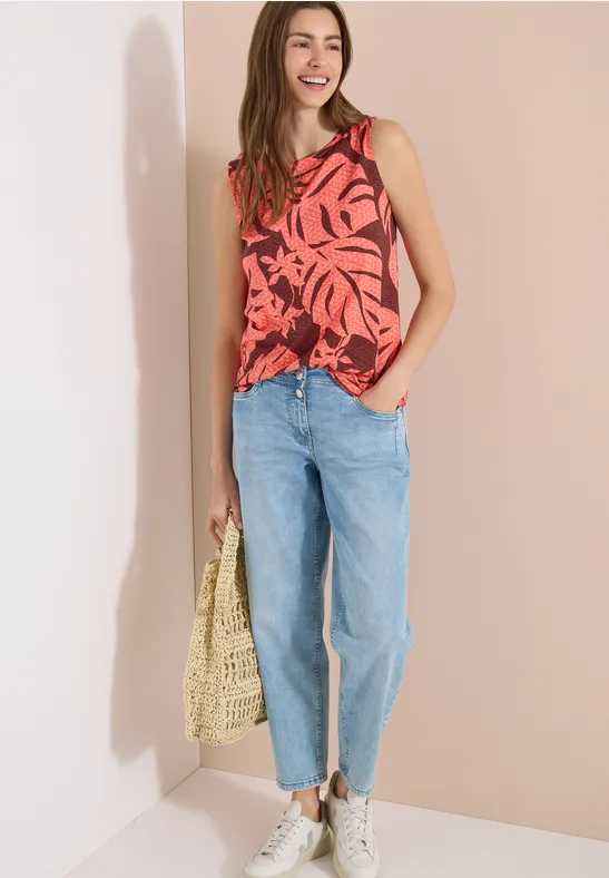 Frau in rotem Top mit Blattmuster, hellblauen Jeans, weißen Sneakers und geflochtener Tasche vor hellem Hintergrund.