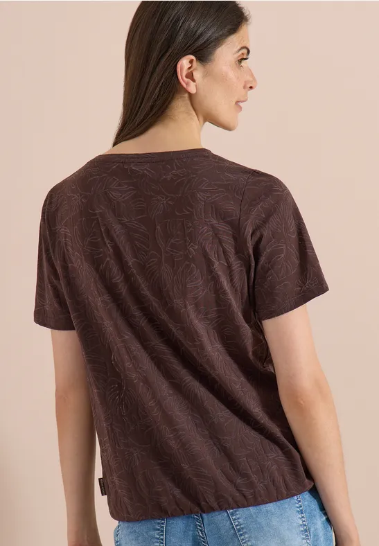 Eine Frau von hinten in einem braunen T-Shirt mit kurzem Arm vor beigem Hintergrund.