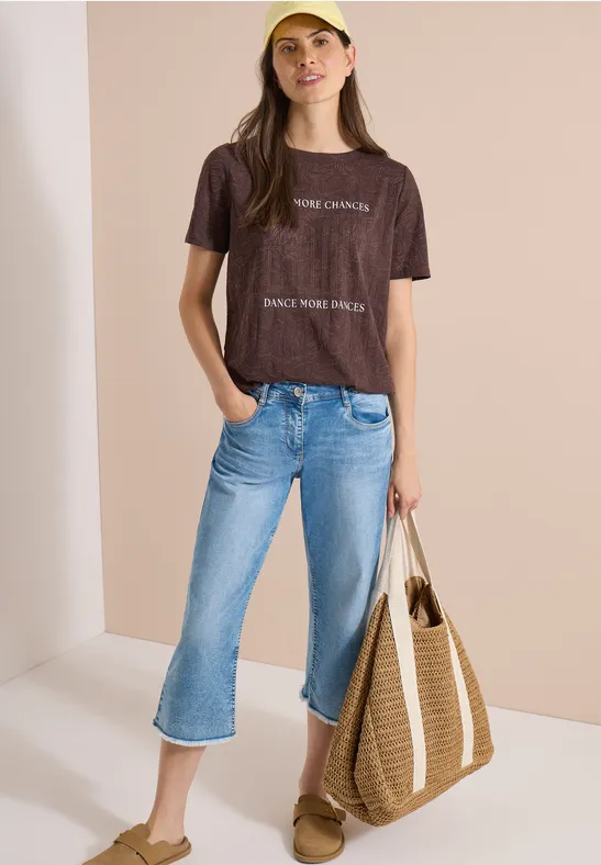 Eine Person trägt braunes T-Shirt, blaue Cropped-Jeans, Sandalen und eine große Strohtasche vor neutralem Hintergrund.
