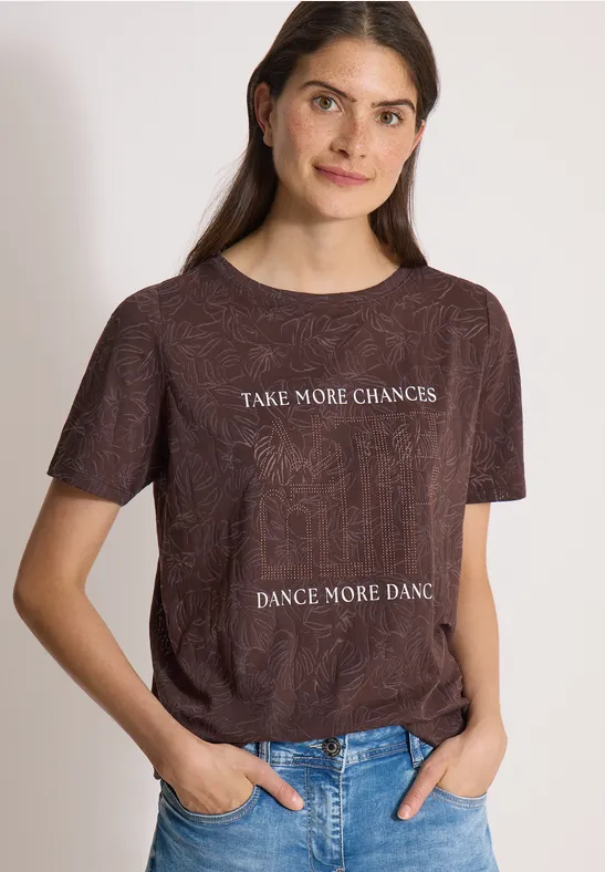 Frau trägt braunes T-Shirt mit weißem Schriftzug 'TAKE MORE CHANCES, DANCE MORE DANCE' und Jeans.