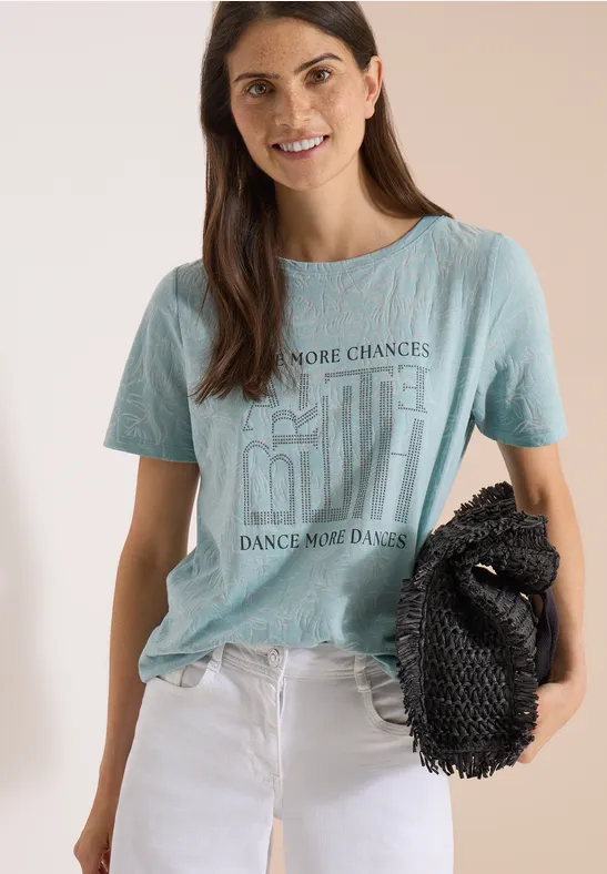 Junge Frau trägt hellblaues T-Shirt mit Aufdruck, weiße Hose und schwarze Clutch, vor neutralem Hintergrund.