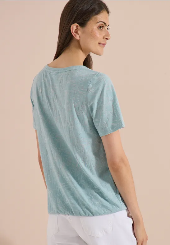 Rückansicht einer Person in grünem T-Shirt und weißer Hose vor neutralem Hintergrund.