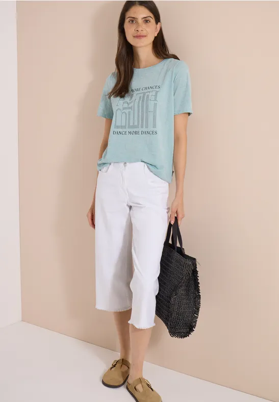 Frau vor beige-eckiger Wand, trägt hellgrünes T-Shirt mit Print, weiße Caprihose, beige Schuhe, schwarze Tasche.