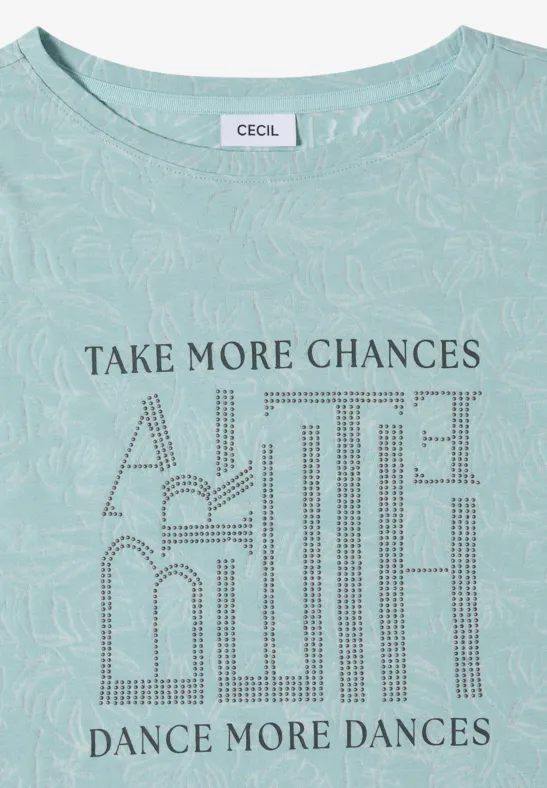 Hellgrünes Shirt mit Blättermuster und der Aufschrift „TAKE MORE CHANCES, DANCE MORE DANCES“.