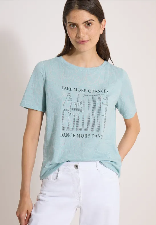 Frau trägt hellgrünes T-Shirt mit Aufschrift und weiße Hose vor hellem Hintergrund.