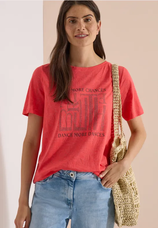 Frau trägt rotes T-Shirt mit Print, blaue Jeans und eine geflochtene Tasche aus Stroh.