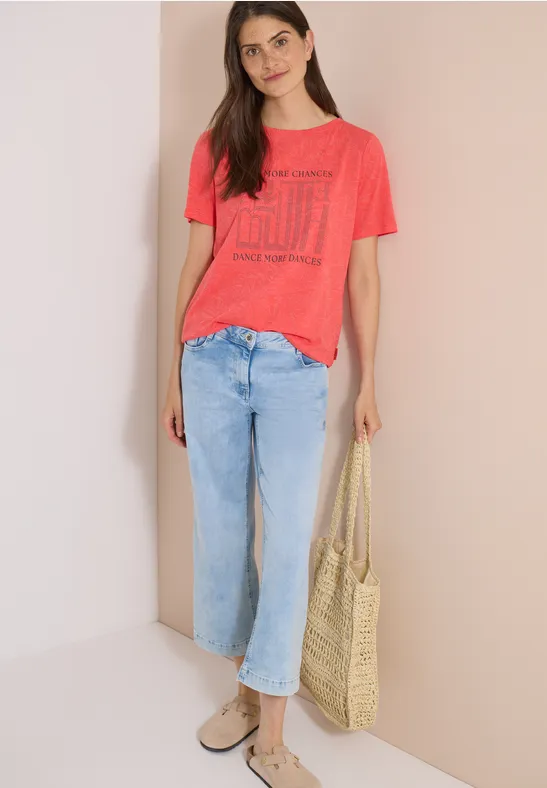 Frau in rotem T-Shirt, hellen Jeans, trägt Basttasche und Sandalen.