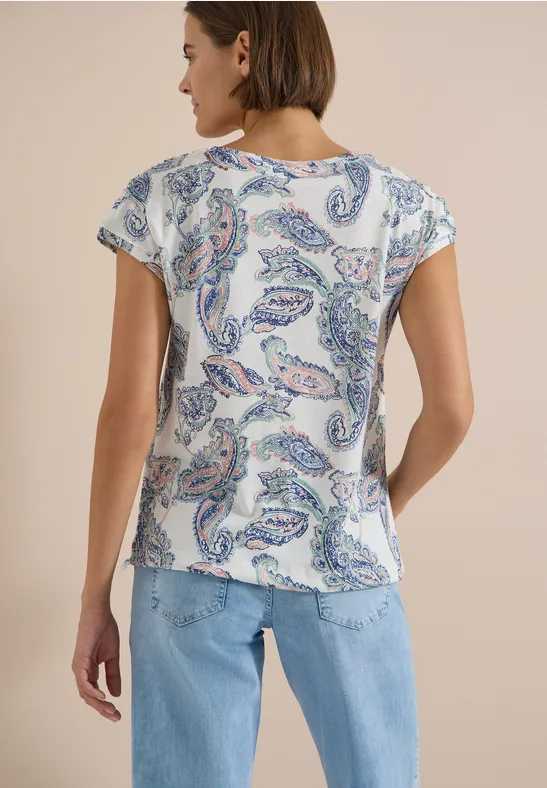 Eine Person von hinten, trägt eine weiße Bluse mit Paisley-Print und blaue Jeans vor beigem Hintergrund.