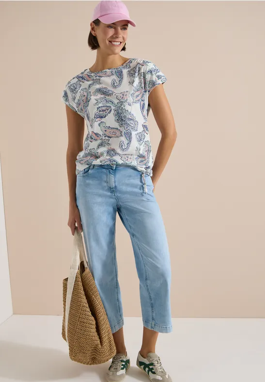 Frau mit gemustertem T-Shirt, hellblauen Cropped-Jeans und Strohtasche vor neutralem Hintergrund.