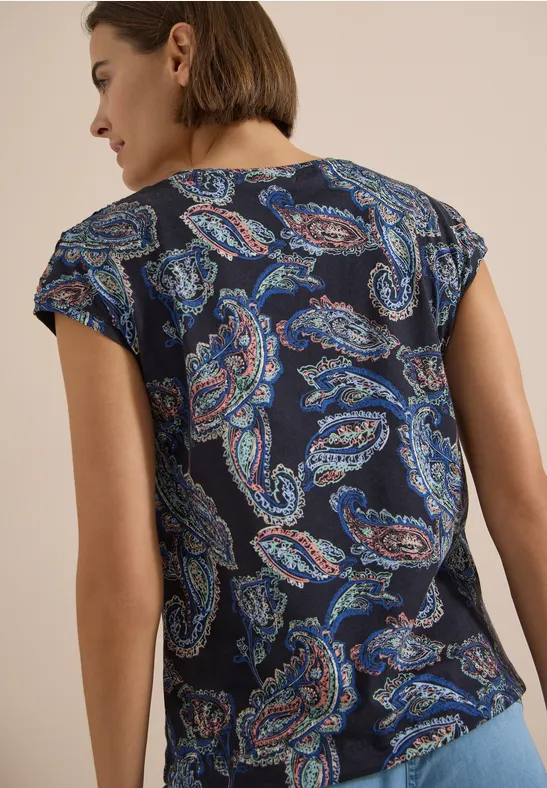 Frau von hinten trägt dunkelblaues T-Shirt mit mehrfarbigem Paisley-Muster, beiger Hintergrund.