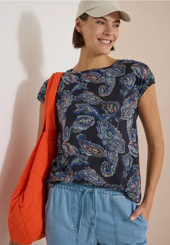 Frau mit paisleygemustertem Shirt, Jeans und roter Schultertasche.