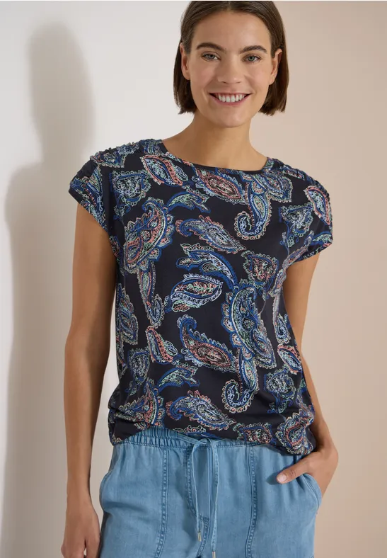 Frau in dunkelblauem T-Shirt mit Paisley-Muster und hellblauer Jeans, lässig mit Händen in den Taschen.