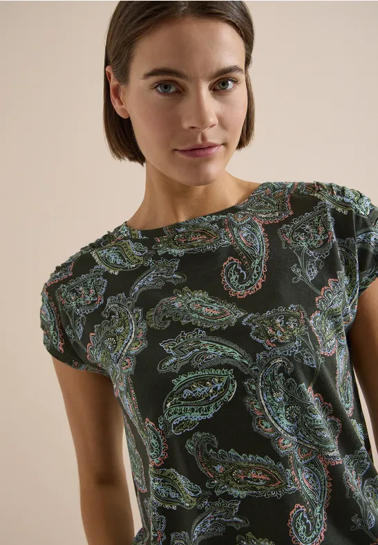 Eine Frau trägt ein dunkelgrünes T-Shirt mit buntem Paisley-Muster vor hellem Hintergrund.