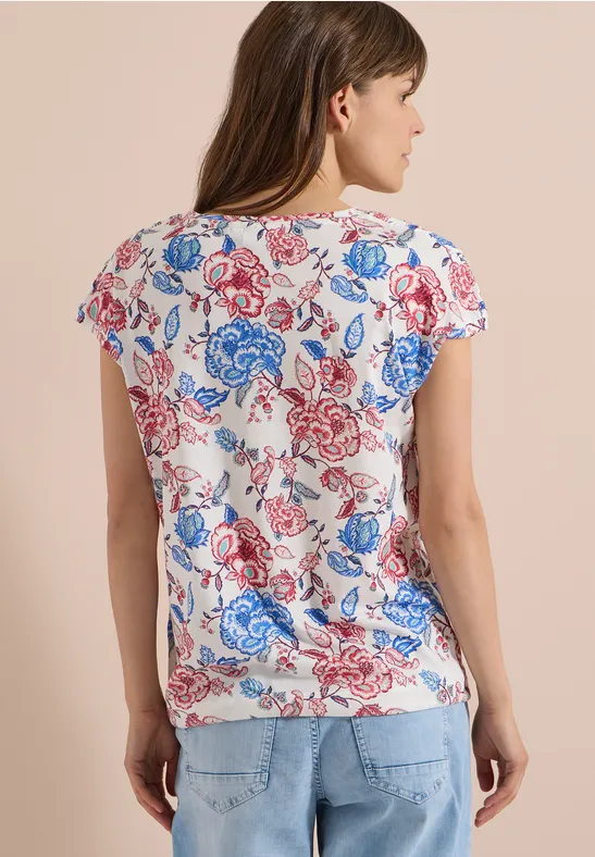 Vrouw van achteren in een wit t-shirt met rood-blauw bloemenpatroon en een spijkerbroek.