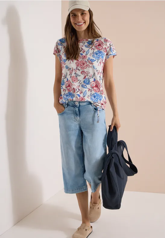 Vrouw draagt bloemen-T-shirt, wijde jeans en houdt een blauwe tas vast.