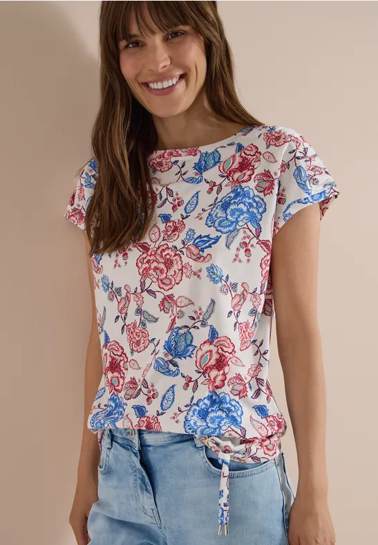 Vrouw met wit T-shirt met blauw-rode bloemenprint en spijkerbroek voor een lichte achtergrond.