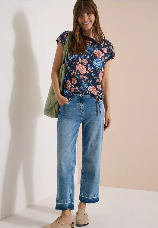 Vrouw met bloemenprint shirt, wijd jeans, beige sandalen en groene tas voor een effen achtergrond.