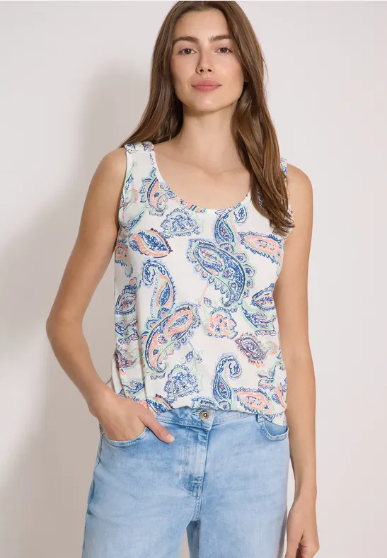 Frau trägt weißes Tanktop mit pastellfarbenem Paisley-Muster und hellblauer Jeans, Hände in den Taschen.