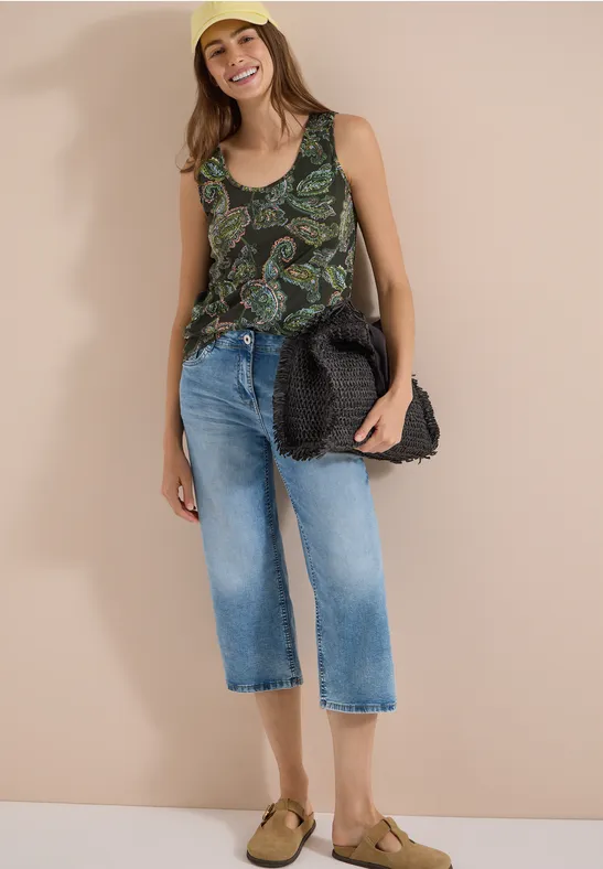 Frau mit unkenntlichem Gesicht in gemustertem Top, Cropped-Jeans und Sandalen mit Strohtasche.