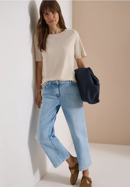 Frau lehnt in heller Jeans und beigem T-Shirt, hält eine dunkelblaue Jacke in der Hand.