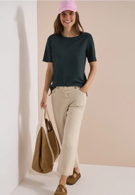 Dame vor beiger Wand in dunkelgrünem T-Shirt, hellen Cordhosen, mit Korbtasche und braunen Sandalen.