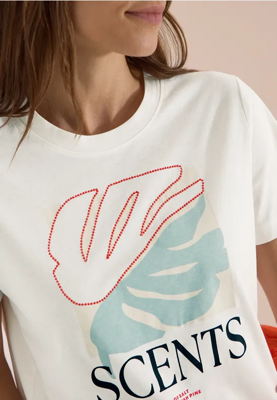 Eine Frau trägt ein weißes T-Shirt mit roten und hellblauen Blattmotiven und dem Schriftzug 'SCENTS'.