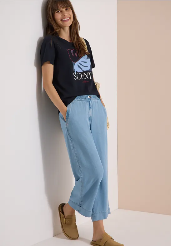 Frau lehnt an Wand, trägt schwarzes T-Shirt mit Print, weite hellblaue Jeans und braune Mokassins.
