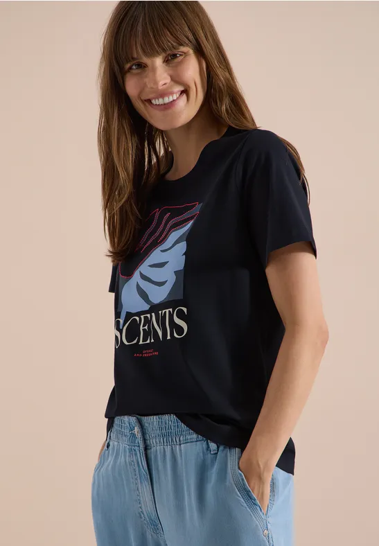 Frau trägt schwarzes T-Shirt mit stilisiertem Gesicht, hellblaue Jeans, steht seitlich mit Hand in Tasche.