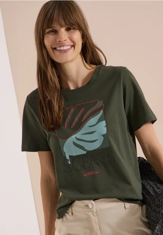 Person trägt ein khakigrünes T-Shirt mit Blättermotiv und eine beige Hose, hält einen dunklen Pullover in der Hand.