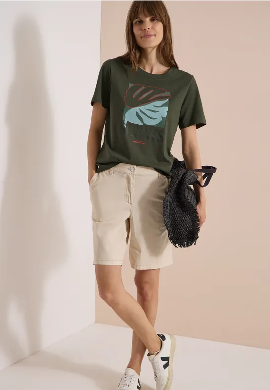 Person lehnt an Wand, trägt grünes T-Shirt mit Blattdesign, beiges Bermuda, weiße Sneaker und schwarzen Netzbeutel.
