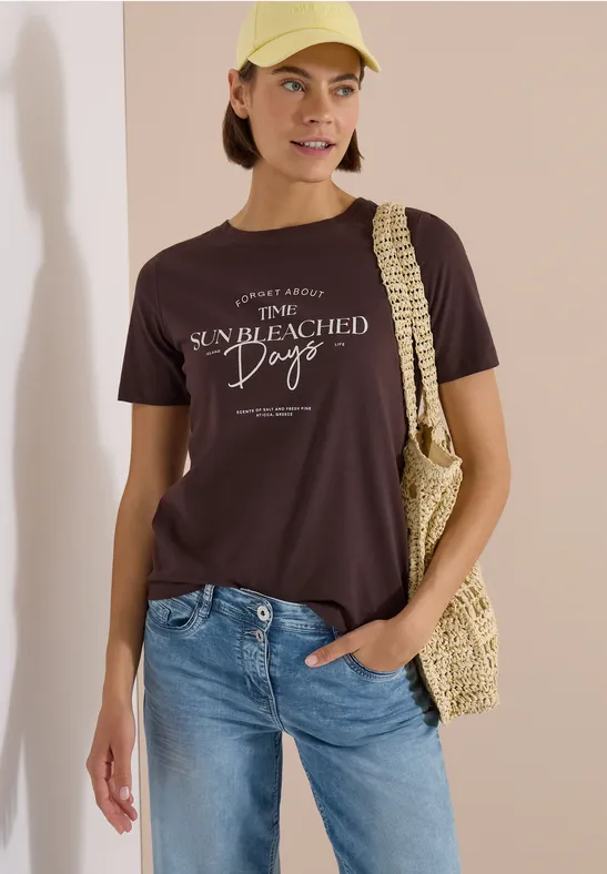 Frau in blauen Jeans und dunkelbraunem T-Shirt mit weißem Print, trägt eine Strohtasche vor hellem Hintergrund.