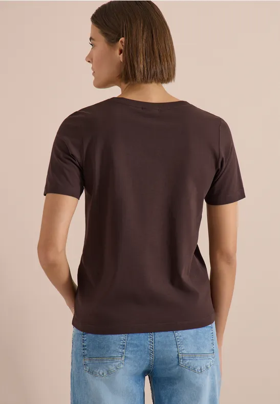 Eine von hinten gesehene Person trägt ein braunes T-Shirt und blaue Jeans vor beigem Hintergrund.