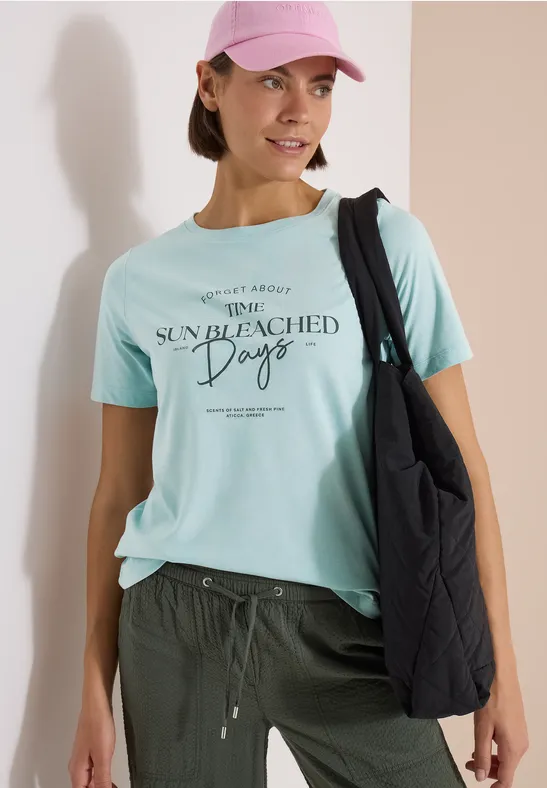 Frau trägt mintfarbenes T-Shirt mit Aufschrift „Sun Bleached Days“ und schwarze Schultertasche.