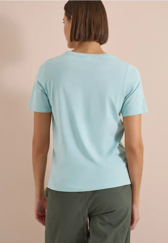 Frau mit kurz geschnittenem Haar trägt ein hellblaues T-Shirt und dunkelgrüne Hose, Ansicht von hinten.