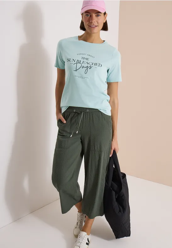 Person trägt mintfarbenes T-Shirt mit Aufdruck, weite grüne Culotte, weiße Sneaker und schwarzen Shopper.