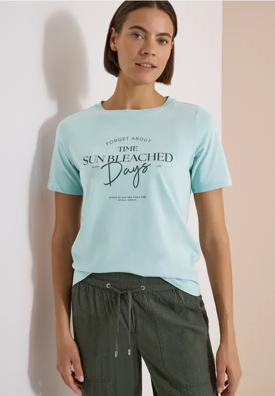 Frau trägt hellblaues T-Shirt mit Aufdruck 'SUN BLEACHED Days' und dunkelgrüne Hose.