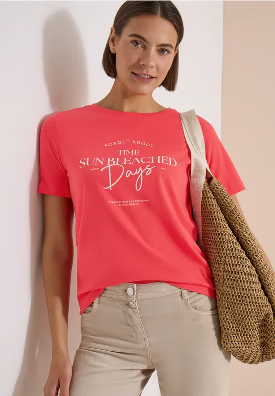 Frau trägt korallrotes T-Shirt mit weißem Aufdruck 'Sun Bleached Days', hellbeige Hose und Strandtasche.
