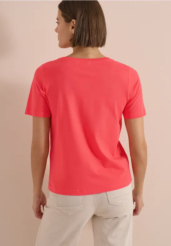 Frau von hinten in korallenrotem T-Shirt und beigen Hosen vor neutralem Hintergrund.