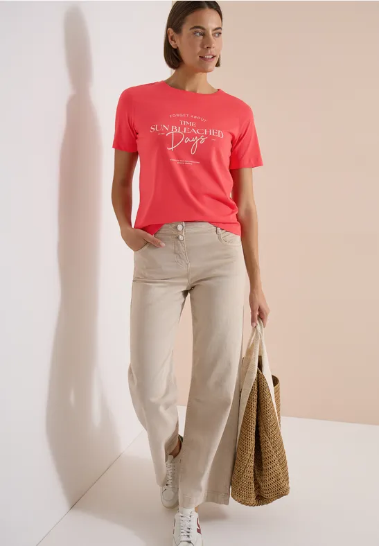 Frau in hellem Korall-T-Shirt, beiger Hose und weißen Sneakern mit großer Basttasche.