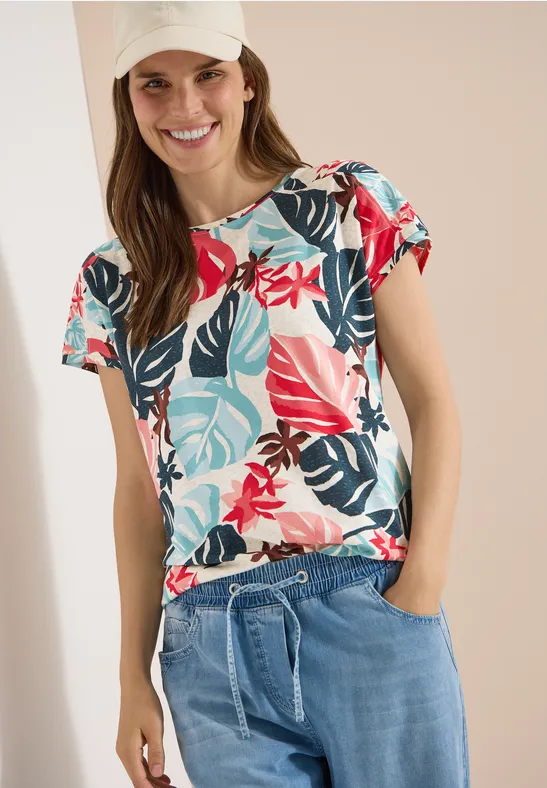 Frau trägt buntes T-Shirt mit tropischem Blumenmuster und leichte Jeanshose mit Kordelzug.