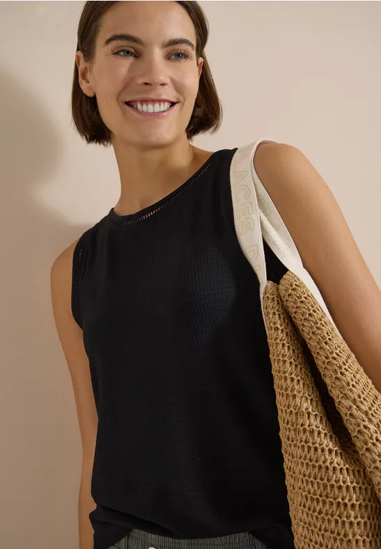 Frau in schwarzem ärmellosem Top mit großem geflochtenem Strohtasche vor neutralem Hintergrund.